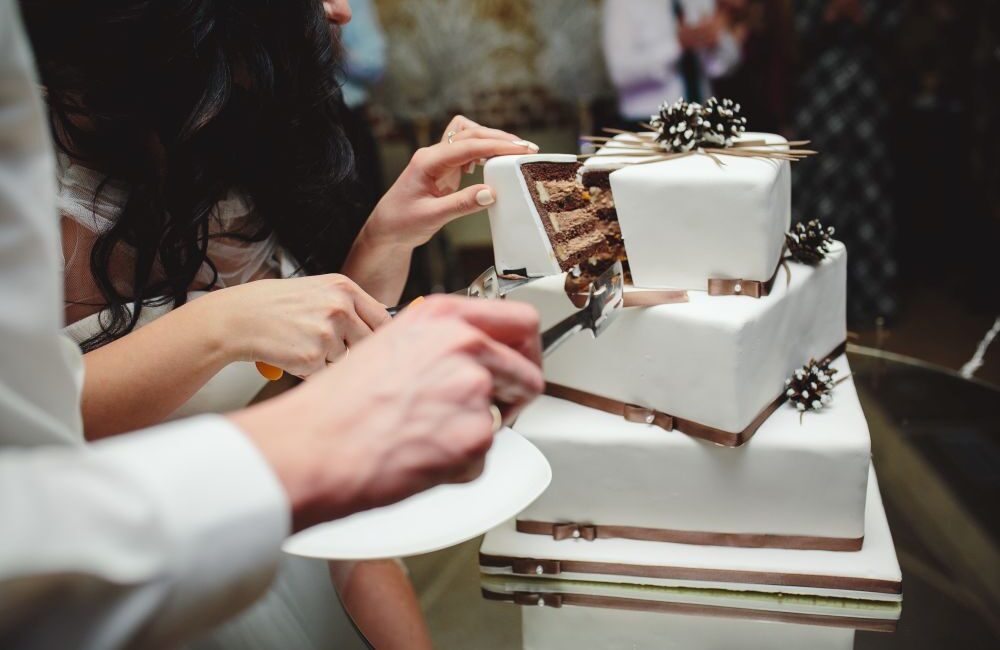 Two people cut a slice from a customised cake Singapore—a three-tiered square white wedding cake with chocolate layers, decorated with brown ribbon and dark flower accents.